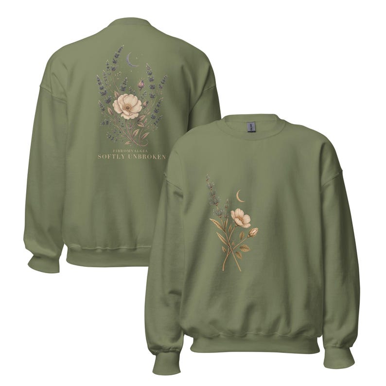 May include: An olive green crewneck sweatshirt featuring a floral graphic with a crescent moon. The back displays a larger bouquet with the text "FIBROMYALGIA SOFTLY UNBROKEN" below it, while the front shows a smaller, matching floral design.