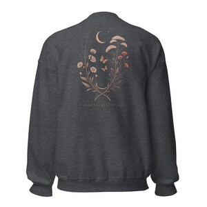 May include: A charcoal grey crewneck sweatshirt featuring a back graphic of a crescent moon, two butterflies, and a floral arrangement of daisies and wildflowers in muted orange and white tones, with the text WALKING BESIDE YOU printed below.