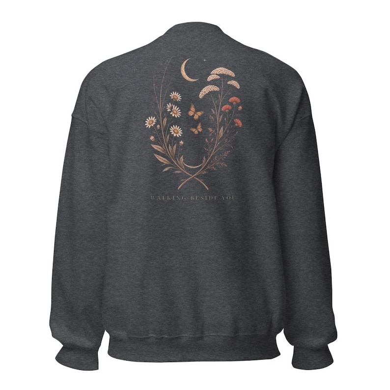 May include: A charcoal grey crewneck sweatshirt featuring a back graphic of a crescent moon, two butterflies, and a floral arrangement of daisies and wildflowers in muted orange and white tones, with the text WALKING BESIDE YOU printed below.