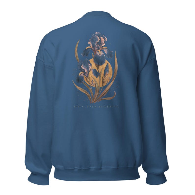May include: A blue crewneck sweatshirt featuring a graphic on the back of two dark purple iris flowers layered over a large golden yellow circle. The text LUPUS - LIVING BEAUTIFULLY is printed in small white letters beneath the floral illustration.