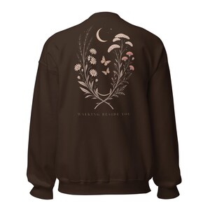 May include: A dark brown crewneck sweatshirt featuring a back graphic of wildflowers, two butterflies, and a crescent moon. The text "WALKING BESIDE YOU" is printed in a light colour below the floral design, which is arranged in a curved shape.