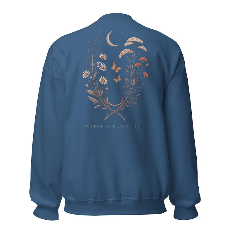 May include: A blue crewneck sweatshirt featuring a back graphic of a crescent moon, two butterflies, and various wildflowers in shades of tan and orange. The text "WALKING BESIDE YOU" is printed in small, light-coloured capital letters below the floral design.