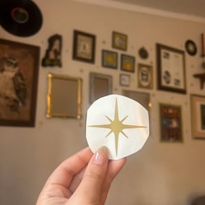 May include: A white sticker with a gold starburst design. The sticker is held in front of a wall decorated with framed pictures and a vinyl record. The starburst has eight points and is centered on the sticker.