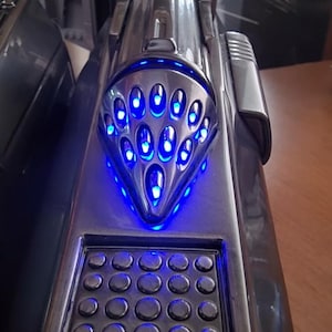 May include: A metallic silver electronic device featuring a pointed, triangular section with multiple oval cutouts illuminated by bright blue LED lights, positioned above a rectangular grid of sixteen small, circular, raised buttons.