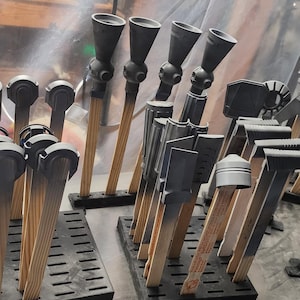 May include: A collection of various black painted objects, possibly parts or components, mounted on wooden dowels and arranged on black slotted trays. The items have a uniform dark color and are in a workshop setting.