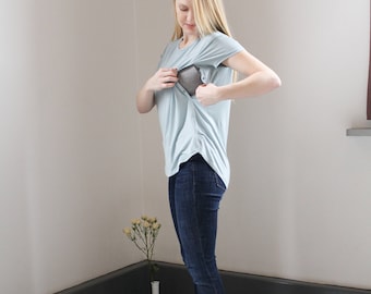May include: A woman wearing a light blue, short-sleeved t-shirt with a built-in nursing panel. She is wearing dark blue jeans and brown ankle boots.