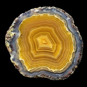 May include: A cross-section of a polished agate stone, showcasing concentric bands of yellow, brown, and gray hues. The center features a geometric pattern, and the outer edge has a rough, textured appearance against a black background. This natural stone is a decorative item.