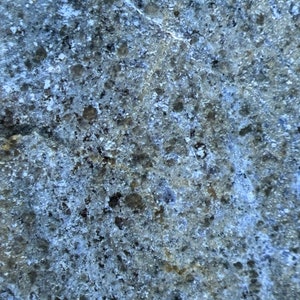 May include: Close-up of a rough, textured surface with a blue-grey color. The surface has a speckled appearance with small, dark grey and white flecks.