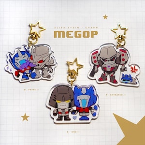 May include: Clear acrylic keychains featuring cartoon versions of Transformers characters. Each keychain has a gold star-shaped clasp. The keychains include characters like Optimus Prime and Megatron, with the text "ALISA AYDIN / CHARM" and "MEGOP".