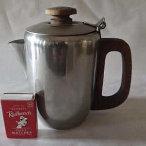May include: A stainless steel percolator coffee pot with a dark brown wooden handle and a matching wooden knob on the lid. A small red box of Redheads Classic Safety Matches is positioned in the foreground to provide a sense of scale.