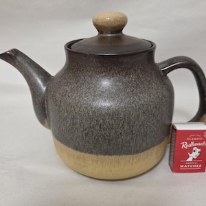 May include: A ceramic teapot with a two-tone glaze featuring a dark brown, speckled upper section and a solid tan base. The teapot has a matching lid with a knob handle and a curved spout. A red Redheads safety matches box is placed beside it.