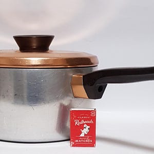 May include: A silver-toned aluminium saucepan with a copper-coloured lid and a dark brown handle. A small red box of Redheads safety matches is placed in front of the pan for scale, showing the relative size of the cookware against the matchbox.