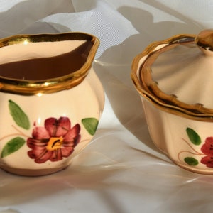 May include: A cream-colored ceramic creamer and sugar bowl set with gold trim and floral designs. The creamer has a handle and the sugar bowl has a lid.