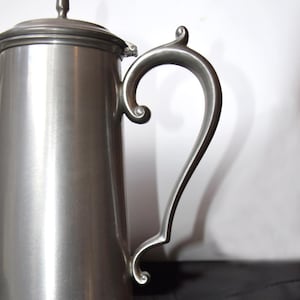 Puede incluir: Una cafetera de metal cepillado con tapa y asa curva. La cafetera tiene un pico y un adorno decorativo en la tapa. La cafetera está sobre una superficie negra.