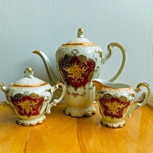 May include: A vintage porcelain tea set with gold and red floral designs. The set includes a teapot, creamer, and sugar bowl.