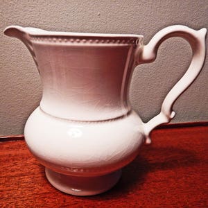 May include: A white ceramic pitcher with a textured surface and a rounded body. The pitcher has a wide, slightly flared spout and a handle that curves up and over the top.