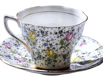 May include: A white teacup and saucer with a floral pattern. The pattern features small black dots, pink, yellow, and blue flowers, and green leaves. The cup and saucer have a gold trim.
