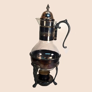 Puede incluir: Una cafetera de plata y vidrio con un asa decorativa y un soporte con tres patas. La cafetera tiene un cuerpo de vidrio transparente y una tapa de plata con un pomo.