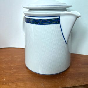 May include: White ceramic coffee pot with a lid. The pot features a blue and green decorative band around the middle and a diamond pattern on the body. The spout is triangular. A classic piece for serving beverages.