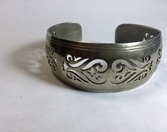 Puede incluir: Un brazalete de metal plateado con un diseño decorativo de corte. El brazalete está abierto y tiene una forma redondeada.
