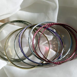 Puede incluir: Un conjunto de brazaletes plateados y dorados con un brazalete rosa y morado. Los brazaletes están apilados uno encima del otro.