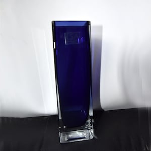 May include: A tall, rectangular cobalt blue glass vase with a clear glass base. The vase has a slightly rounded shape and is made of thick glass. The vase is empty and has a clear glass base.