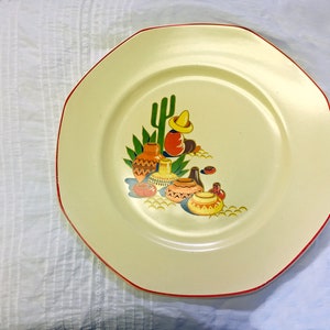Puede incluir: Un plato de cerámica blanco con un borde rojo presenta una ilustración colorida de un cactus, cerámica y una persona con un sombrero. El diseño es de estilo suroeste.