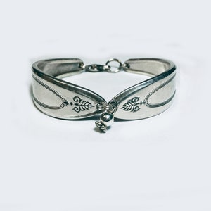 Puede incluir: Un brazalete de plata hecho de dos mitades de cuchara, conectadas por una cadena y una pequeña perla de plata.