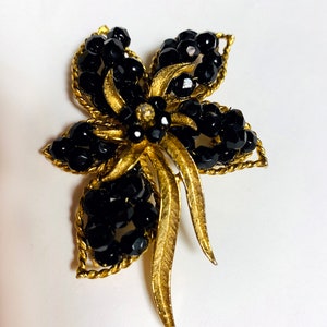Puede incluir: Broche floral vintage con marco de metal dorado y detalles de cuentas negras. El broche presenta un diseño floral con múltiples pétalos, cada uno adornado con cuentas negras. El centro de la flor tiene un pequeño adorno brillante.