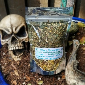 May include: A clear plastic bag filled with dried herbs and labeled "Deez Bonez." The label lists ingredients including nettle, peppermint, and licorice. A decorative skull is in the background, suggesting a theme of herbal remedies.