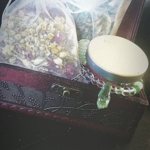 May include: A wooden box with a hinged lid, filled with various items. The box is decorated with a floral pattern and has a metal latch. Inside the box, there is a white mesh bag filled with dried herbs, a small glass jar with a lid, and a beaded necklace with green and clear beads.