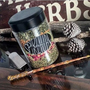 Könnte beinhalten: Ein Glasgefäß gefüllt mit getrockneten Kräutern und Blumen, beschriftet mit "Spiritual  Badass  Ritual". Das Gefäß steht auf einer Holzoberfläche mit einem Holzstab und Tannenzapfen.