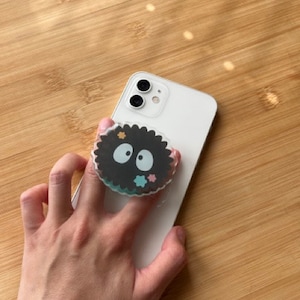 Peut inclure: Un iPhone blanc avec une poignée de téléphone noir et blanc représentant un personnage de dessin animé avec un corps noir et des yeux blancs. La poignée de téléphone est fixée à l'arrière du téléphone.