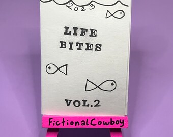 Könnte beinhalten: Ein weißes Heftchen mit dem Titel "Februar 2023 Life Bites Vol. 2" und einer Zeichnung von Fischen. Das Heftchen wird auf einem rosafarbenen Staffelei mit der Aufschrift "Fictional Cowboy" präsentiert.