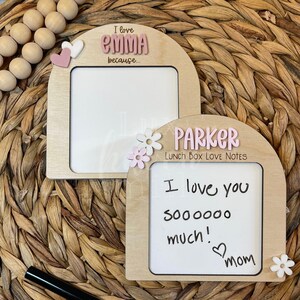 May include: Two wooden frames with white dry-erase surfaces. The frames are shaped like arches and have pink and white flowers and hearts attached. The top frame says "I love Emma because..." and the bottom frame says "Parker Lunch Box Love Notes" and has the message "I love you sooooooo much! ♡ mom" written on it.