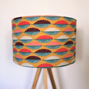 Peut inclure: Abat-jour jaune moutarde avec un motif répété de formes de feuilles stylisées en teal, rouge, blanc, rose et marron. L'abat-jour est cylindrique et repose sur un pied en bois.