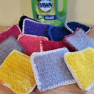 May include: A stack of colorful crocheted dishcloths with a bottle of Dawn Ultra Antibacterial dish soap in the background. The dishcloths are made with a sparkly yarn and have a white crocheted border.