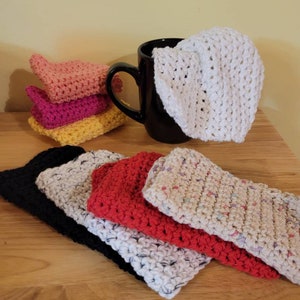 May include: A collection of handmade crochet washcloths in various colors, including pink, yellow, black, white, and red. A black mug holds a white washcloth. The washcloths are stacked and arranged on a wooden surface.