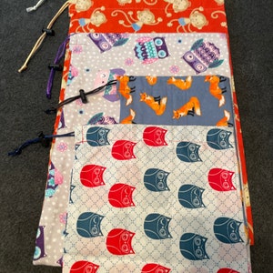 May include: Five reusable fabric bags with different colorful patterns. The bags are stacked on top of each other. The top bag has a green background with a white star pattern. The second bag has an orange background with a monkey pattern. The third bag has a gray background with an owl pattern. The fourth bag has a blue background with a fox pattern. The bottom bag has a white background with a red and blue owl pattern.