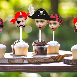 May include: Five cupcakes with pirate themed toppers featuring children's faces. The toppers are red, black, and white with a pirate hat, eye patch, and bandana.