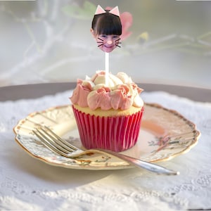 Puede incluir: Un cupcake rosa y blanco con un forro de cupcake rojo. La cara de una niña con un filtro de cara de gato está en un palo en el cupcake.