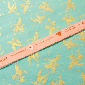 Peut inclure: Gros plan sur un tissu turquoise avec un motif répété de poissons dorés. Une règle rose avec le texte "RUBY STAR SOCIETY" et "missmelodymiller" est en diagonale sur le tissu. Le tissu est 100% coton et fabriqué au Japon.