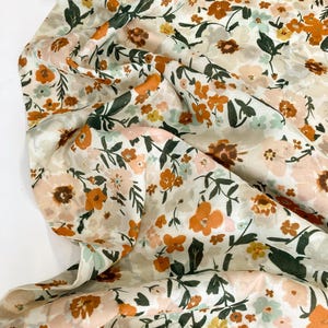 May include: Cream-coloured floral fabric featuring a pattern of orange, pink, yellow, and light blue flowers. Dark green leaves and stems are scattered throughout the design. The fabric is folded, creating shadows and depth.