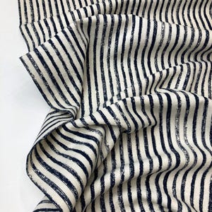 May include: Close-up of a fabric with a repeating pattern of navy blue vertical stripes on an off-white background. The fabric is folded and draped, showing the texture and direction of the stripes.