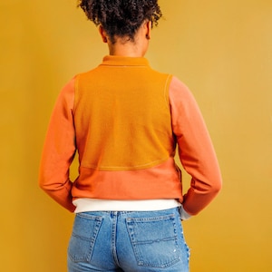 Puede incluir: Una persona lleva una camisa de manga larga con un dise&ntilde;o de bloques de color marr&oacute;n y naranja. La camisa tiene un cuello blanco y una capa inferior blanca. La persona lleva pantalones vaqueros azules.