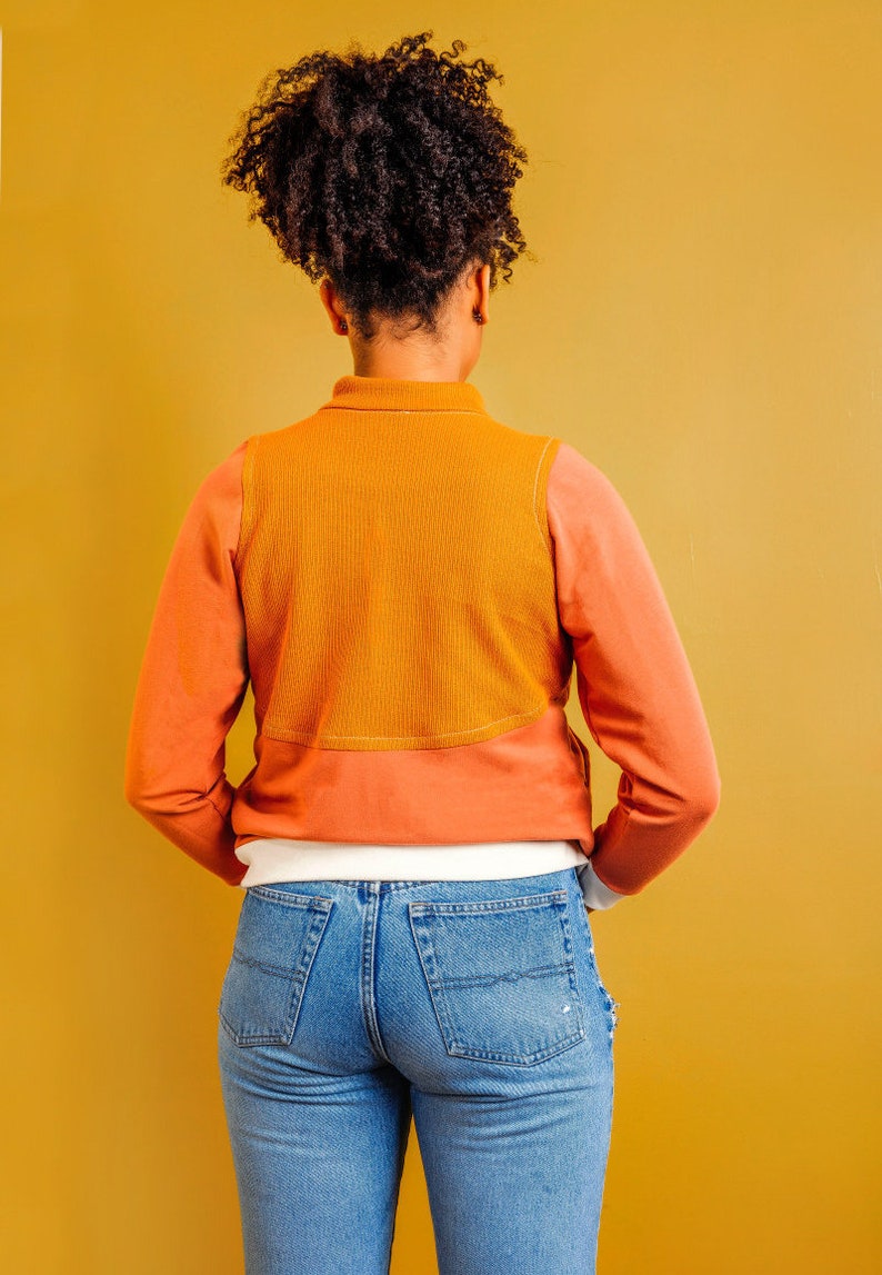 Puede incluir: Una persona lleva una camisa de manga larga con un dise&ntilde;o de bloques de color marr&oacute;n y naranja. La camisa tiene un cuello blanco y una capa inferior blanca. La persona lleva pantalones vaqueros azules.