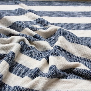 May include: A white and blue striped throw blanket with a textured weave. The blanket is folded and draped over itself, showing the stripes and texture.