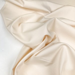 May include: Cream-colored fabric with soft folds and a smooth texture. The fabric is draped, creating shadows and highlights that emphasize its flow and drape. Ideal for apparel or home decor.