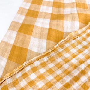 May include: Mustard yellow and white checked fabric. The fabric is folded, displaying the pattern in detail. The edges of the fabric are frayed, adding a rustic touch. The pattern is a classic gingham design.