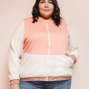 Puede incluir: Una mujer lleva una chaqueta con cremallera rosa y blanca con un ribete de estampado floral. La chaqueta tiene una cremallera blanca y un bolsillo de parche en el lado izquierdo. Lleva pantalones vaqueros azules.